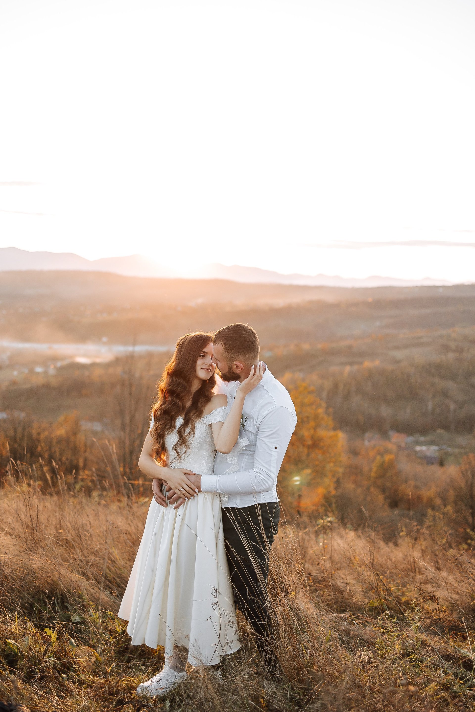 A couple is standing on a hillside, with the sun setting behind them. They are embracing each other, and the mood of the image is romantic and intimate