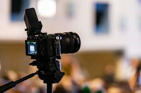 Professional camera on tripod recording a seminar with blurred audience in background. Videography equipment for live event coverage.
