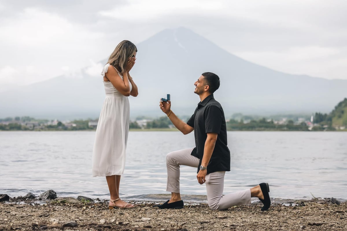 Man proposing to woman on beach with mountain lake backdrop, woman in white dress covering mouth in surprise