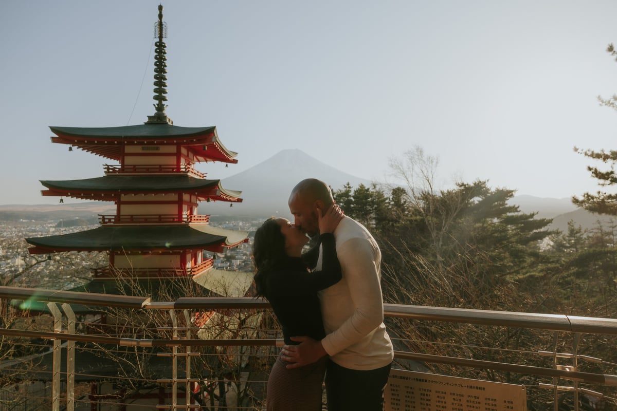 Couple kissing on a wooden railing with a red and green pagoda and Mt. Fuji in the background at an observation point