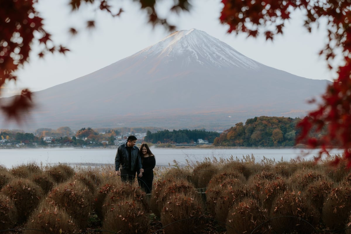 Two people standing in dried grass with Mount Fuji snow-capped peak in the background across a lake