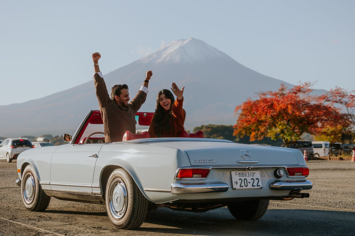 White vintage convertible car with people celebrating inside, Mount Fuji in background, autumn foliage visible