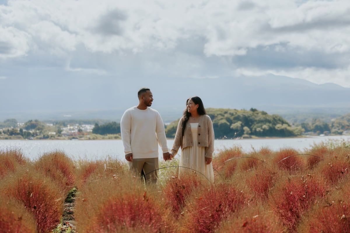 Couple holding hands standing in a field of red flowering plants with a lake and mountains in the background