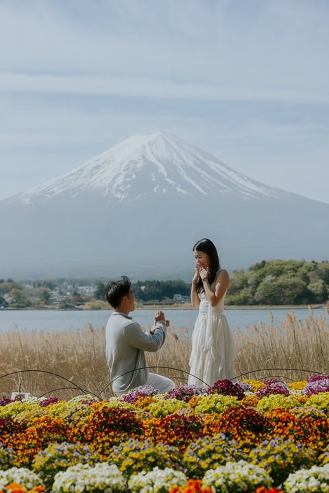 Man proposing to woman in white dress with colorful flowers, lake, and snow-capped Mount Fuji in background