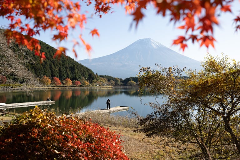 Mount Fuji reflected in a calm lake surrounded by autumn foliage and a person standing on a wooden dock