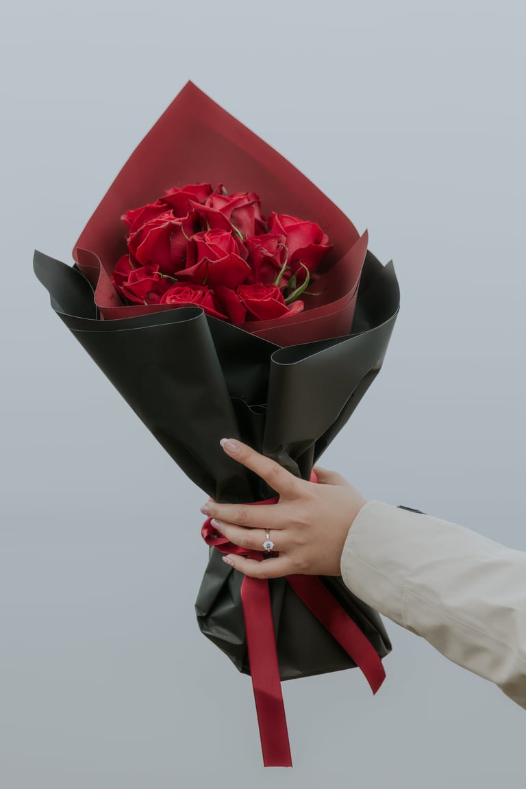 Hand holding a bouquet of red roses wrapped in black paper with a red ribbon against a gray background