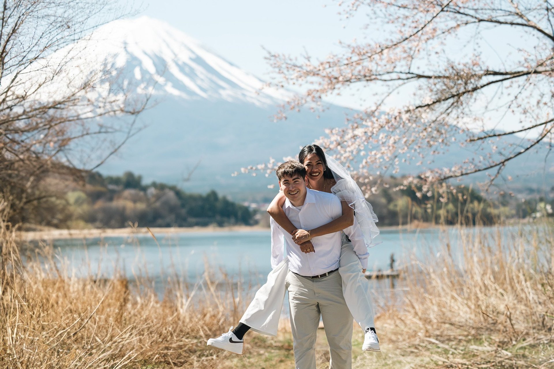 Couple playing piggyback in field with snow-capped Mount Fuji and lake in background