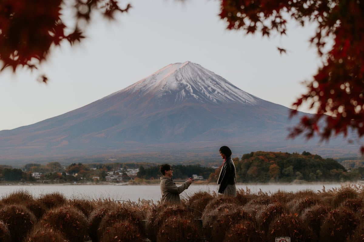 Two people standing by a lake with snow-capped Mount Fuji in the background, framed by red maple leaves in foreground